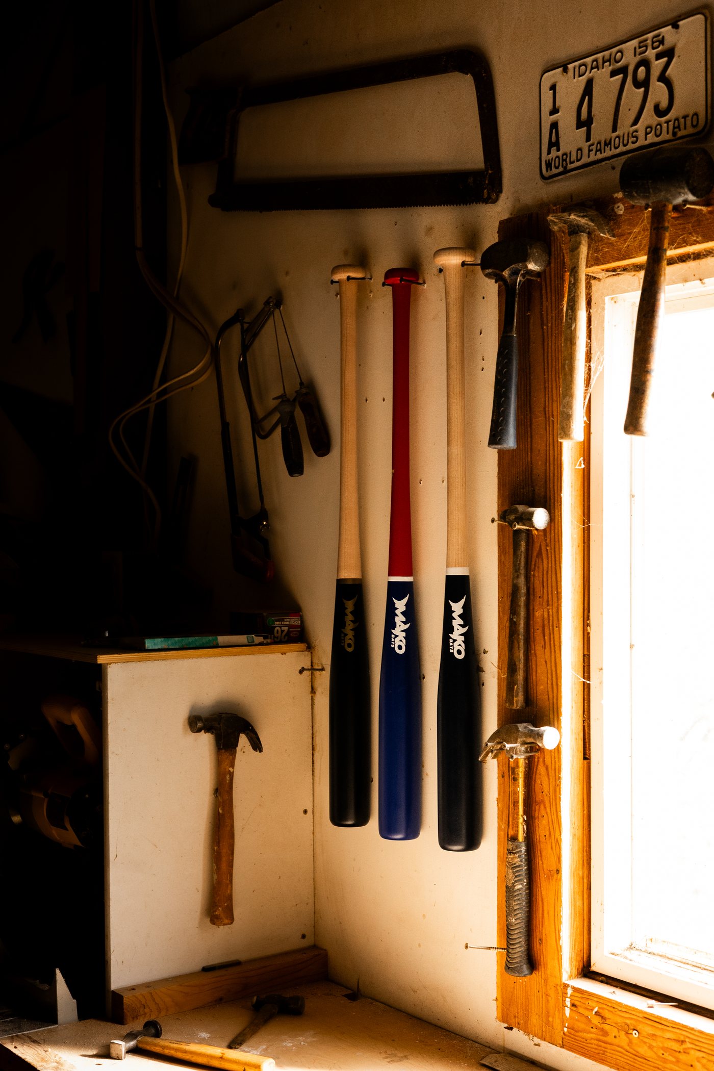 Three Mako bats hanging on the wall of the woodshop beside hand tools, lit by a window.